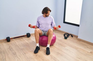 Spor merkezinde topun üzerine oturan halterli orta yaşlı adam eğitimi.