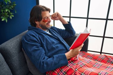 Göz bandı takan orta yaşlı bir adam evdeki kanepede kitap okuyor.