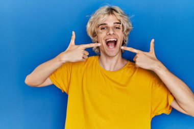 Mavi arka planda duran genç, neşeli bir şekilde gülümseyen ve parmaklarıyla ve ağzıyla işaret eden modern bir adam. diş sağlığı kavramı. 