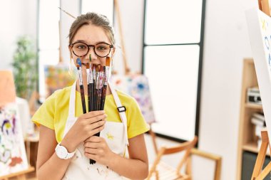 Adorable girl covering mouth with paintbrushes at art studio