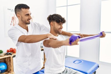 Two hispanic men physiotherapist and patient having rehab session using elastic band at clinic