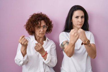 İspanyol anne ve kızı pembe arka plan üzerine gündelik beyaz tişört giyiyorlar. Yumruk savunma hareketleriyle kavga etmeye hazırlar. Kızgın ve üzgün bir yüz. 