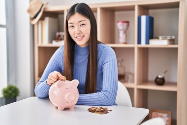 Çinli genç bir kadın domuz kumbarasına para koyuyor. Olumlu ve mutlu görünüyor. Kendine güvenen bir gülümsemeyle dişlerini gösteriyor. 