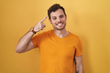 Sarı arka planda duran İspanyol genç adam gülümseyerek tek parmağıyla kafasını işaret ediyor, harika bir fikir ya da düşünce, iyi bir hafıza. 