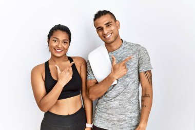 Spor kıyafetleri giyen genç bir çift izole edilmiş arka planda neşeli bir şekilde duruyorlar. Yüzlerinde mutlu ve doğal bir ifade olan elleri ve parmaklarıyla gülümsüyorlar. 