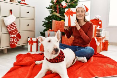 Kafkasyalı genç bir kadın elinde hediyeyle evde köpekle oturuyor.