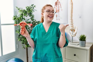 Genç kızıl saçlı kadın, kadın üreme organının anatomik modelini tutuyor. Parmağını yan tarafa uzatıyor ve ağzı açık gülümsüyor. 