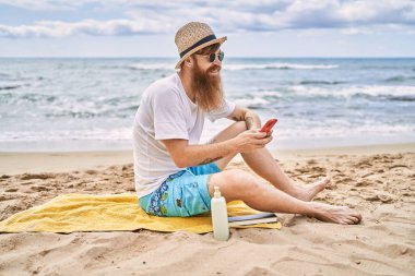Genç kızıl saçlı adam kumsalda havluya oturmuş akıllı telefon kullanıyor..