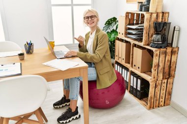 Ofiste çalışan beyaz, güzel bir iş kadını pilates topunun üzerine oturmuş başparmağını yana uzatıyor ve ağzı açık bir şekilde gülümsüyor. 