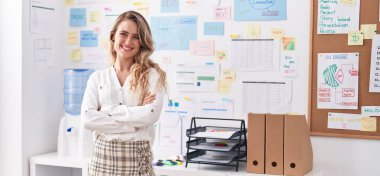 Genç, sarışın, iş kadını, gülümseyen, kendine güvenen, kollarını kavuşturup ofiste el kol kola duran.