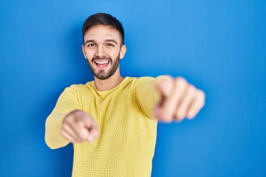 Mavi arka planda duran İspanyol adam sana ve kameraya parmaklarıyla bakıyor, gülümsüyor, pozitif ve neşeli. 