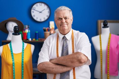 Orta yaşlı, gri saçlı, sırıtan, kendine güvenen terzi terzide kollarını kavuşturup el kol hareketi yapan bir adam.