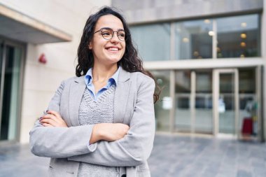 Genç İspanyol kadın yönetici gülümsüyor. Kollarını kavuşturup caddede duruyor.