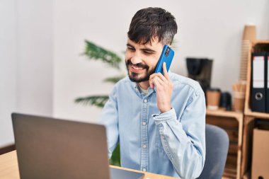 Genç İspanyol iş adamı ofisteki bilgisayarla akıllı telefondan konuşuyor.
