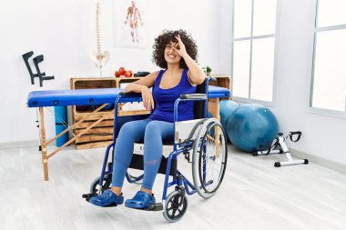 Orta Doğulu genç bir kadın, fizyoterapi kliniğinde tekerlekli sandalyede oturuyor. El hareketleriyle gülümsüyor ve mutlu yüzüyle parmaklarının arasından bakıyor.. 