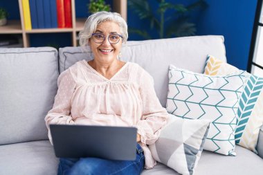 Evdeki kanepede oturan orta yaşlı, kır saçlı bir kadın dizüstü bilgisayar kullanıyor.
