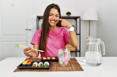 Genç esmer kadın suşi yerken yemek çubuklarıyla parmaklarını yüzüne ve burnuna doğrultarak neşeli bir şekilde gülümsüyor. güzellik konsepti 