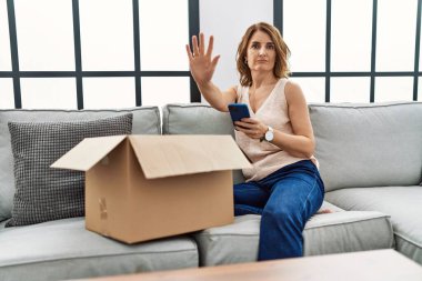 Orta yaşlı bir kadın kanepede oturmuş telefonda teslimat paketini kontrol ediyor. Elinde ciddi ve kendinden emin ifadesiyle dur işareti yapıyor. Savunma jesti. 