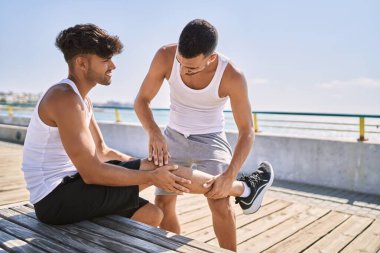 İki İspanyol erkek, deniz kenarında bacağından sakatlanmak için yedek kulübesinde oturuyor.