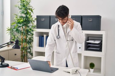 Doktor üniforması giyen İspanyol genç adam klinikte dizüstü bilgisayarla telefonla konuşuyor.