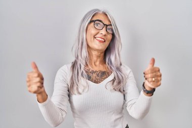 Beyaz arka planda beyaz saçlı, orta yaşlı bir kadın elinde olumlu bir jest yapıyor, gülümsüyor ve mutlu. Neşeli ifade ve kazanan jest. 