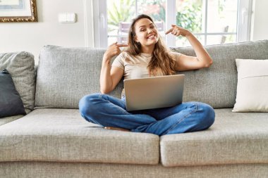 Güzel, genç, esmer bir kadın evdeki bilgisayar laptopunu kullanarak koltukta oturuyor neşeli bir şekilde gülümsüyor ve parmaklarıyla ve ağzıyla işaret ediyor. diş sağlığı kavramı. 