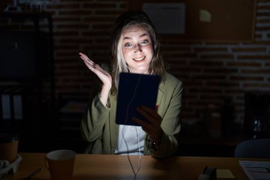Geceleri ofiste çalışan sarışın beyaz bir kadın neşeli bir şekilde gülümsüyor ve avuç içi kameraya bakıyor.. 