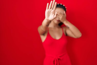 Latin kökenli genç bir kadın kırmızı arka planda duruyor. Gözleri ellerle kaplıyor ve üzgün ve korku dolu bir ifadeyle dur hareketi yapıyor. utangaç ve olumsuz kavram. 