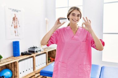 Ağrı kesici kliniğinde çalışan genç bir fizyoterapist kadının elinde kredi kartı var. Parmaklarıyla imza atıyor, gülümseyerek mükemmel bir sembol oluşturuyor. 