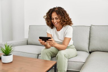 Orta yaşlı İspanyol kadın evde akıllı telefon kullanarak gülümsüyor.