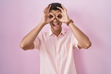 Pembe arka planda duran genç bir İspanyol dürbün gibi jest yapıyor, gözleri parmaklarından bakıyor. çılgın ifade. 