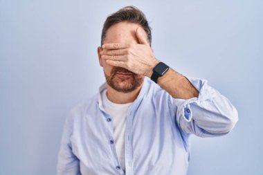 Orta yaşlı, beyaz bir adam mavi arka planda dikiliyor, gözlerini eliyle kapatıyor, ciddi ve üzgün görünüyor. Görme, saklama ve reddetme konsepti 