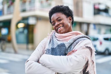 Afro-Amerikalı kadın gülümsüyor. Kollarını kavuşturup caddede duruyor.