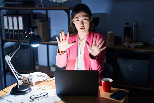 Chinese young woman working at the office at night afraid and terrified with fear expression stop gesture with hands, shouting in shock. panic concept.