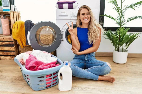 Young beautiful woman doing laundry sitting by wicker basket smiling with happy face winking at the camera doing victory sign. number two.