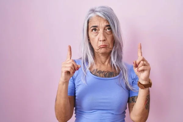 有纹身的中年妇女站在粉红的背景上 满脸愁容 用手指指示方向 忧郁而不快乐 — 图库照片