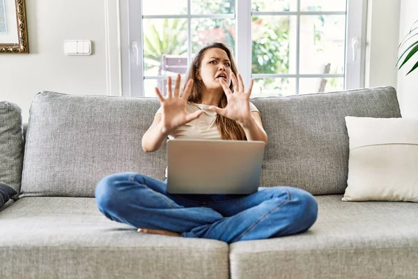 Güzel, genç, esmer bir kadın evdeki bilgisayar laptopunu kullanarak koltukta oturuyor. Korkudan dehşete düşmüş ve korkmuş bir şekilde. El hareketlerini durdur, şok içinde bağır. panik konsepti. 
