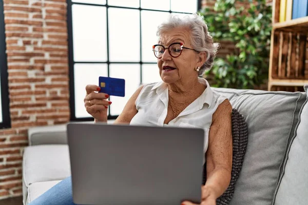 Senior grey-haired woman using laptop and credit card sitting on sofa at home