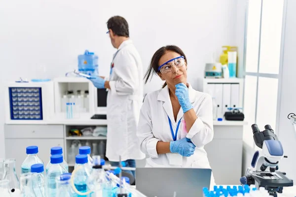 Middle age woman working at scientist laboratory serious face thinking about question with hand on chin, thoughtful about confusing idea