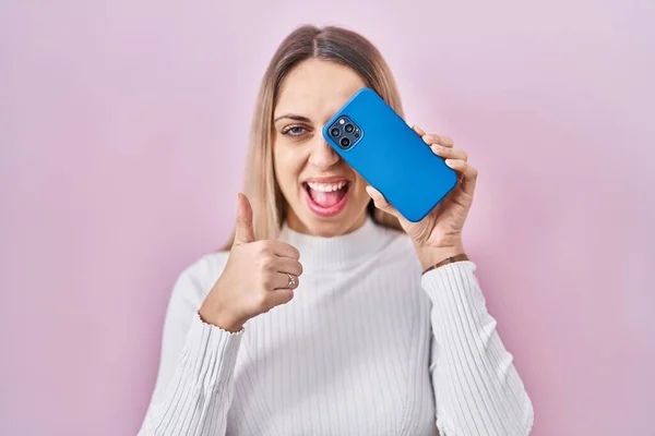 Młoda Blondynka Trzyma Smartfon Okrywający Oko Uśmiechnięty Szczęśliwy Pozytywny Kciuk — Zdjęcie stockowe