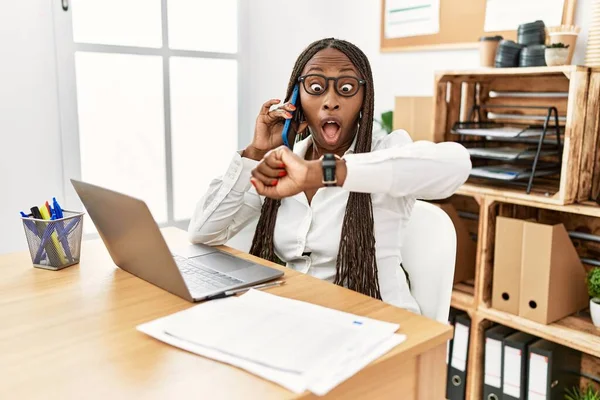 Black woman with braids working at the office speaking on the phone looking at the watch time worried, afraid of getting late