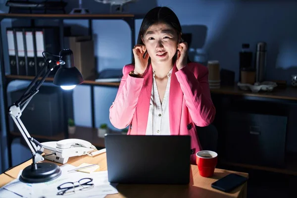 Chinese young woman working at the office at night covering ears with fingers with annoyed expression for the noise of loud music. deaf concept.