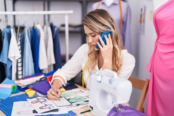 Young blonde woman tailor talking on smartphone writing on notebook at tailor shop