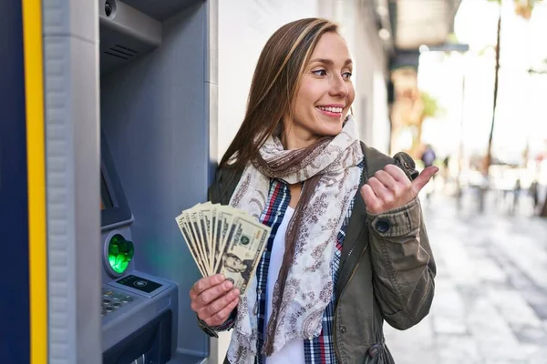 Young Blonde Woman Holding Dollars Banknotes Atm Machine Pointing Thumb — Stock Photo, Image