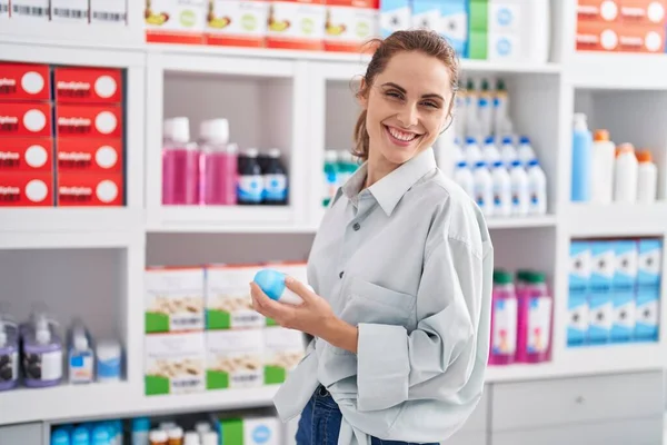 Young woman customer smiling confident holding deodorant bottles at pharmacy