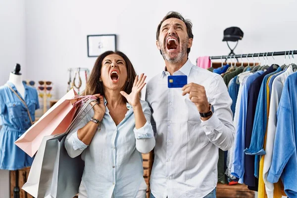 Hispanic middle age couple holding shopping bags and credit card crazy and mad shouting and yelling with aggressive expression and arms raised. frustration concept.