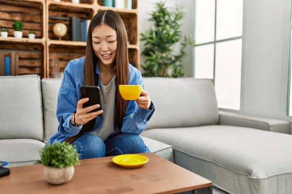 Giovane Ragazza Cinese Utilizzando Smartphone Bere Caffè Seduto Sul Divano — Foto Stock