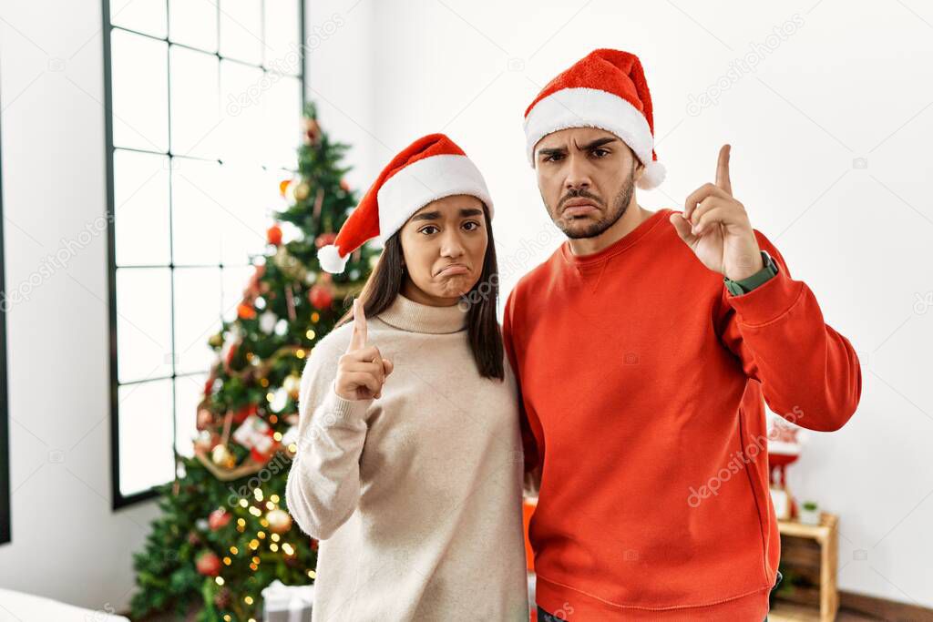 Young hispanic couple standing by christmas tree pointing up looking sad and upset, indicating direction with fingers, unhappy and depressed. 