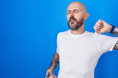 Mavi arka planda dikilen İspanyol dövmeli adam geriliyor, yorgun ve rahat, uykulu ve sabahın erken saatlerinde esniyor. 