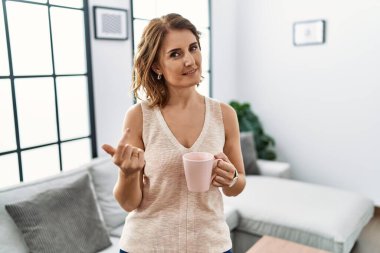 Orta yaşlı bir kadın evde kahve içiyor. Buraya el kol hareketi yaparak mutlu ve gülümseyen birini davet ediyor. 